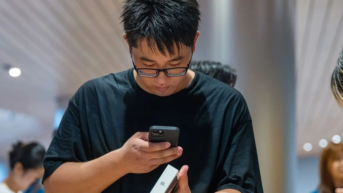 Person in schwarzem T-Shirt mit Brille prüft schwarzes Smartphone und weiße Box. Hintergrund zeigt verschwommene Personen und Lichter.