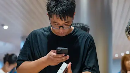 Person in schwarzem T-Shirt mit Brille prüft schwarzes Smartphone und weiße Box. Hintergrund zeigt verschwommene Personen und Lichter.