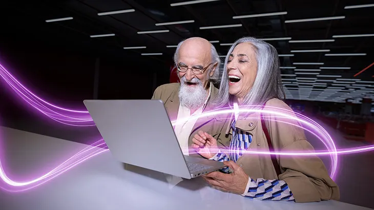 Älteres Paar sitzt lachend vor einem Laptop; leuchtende violette Linien symbolisieren digitale Vernetzung in moderner Umgebung.