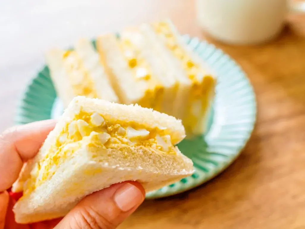 Hand hält dreieckiges Eiersalatsandwich mit Weißbrot. Weitere Sandwiches auf grünem Teller; Holztisch im Hintergrund.