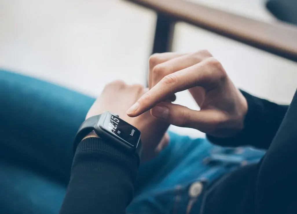 Eine Hand mit dunkler Smartwatch zeigt "12:15 PM Tuesday". Ein Finger tippt auf den Bildschirm. Blaue Jeans und ein Holzstuhl sind im Hintergrund.