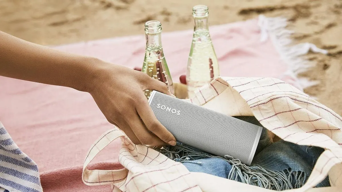 Eine Hand legt einen weißen Sonos Lautsprecher in eine Tasche am Strand.