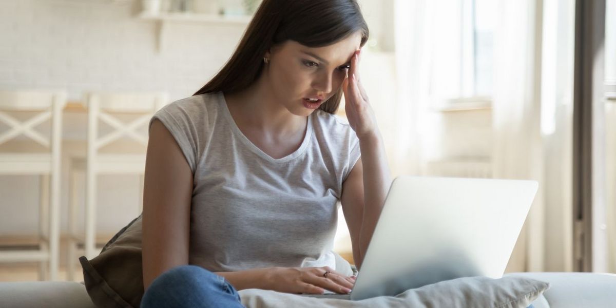 Genervte Frau in grauem Shirt hält Hand an Stirn vor Laptop.