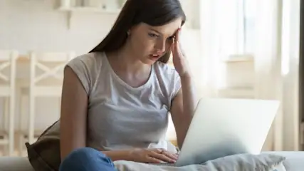 Genervte Frau in grauem Shirt hält Hand an Stirn vor Laptop.