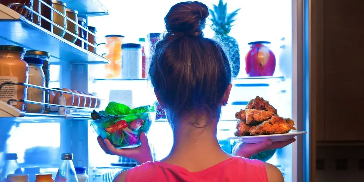 Person von hinten in Pink hält Salat und Brathähnchen vor offenem, beleuchtetem Kühlschrank mit Gläsern, Eiern, Ananas.