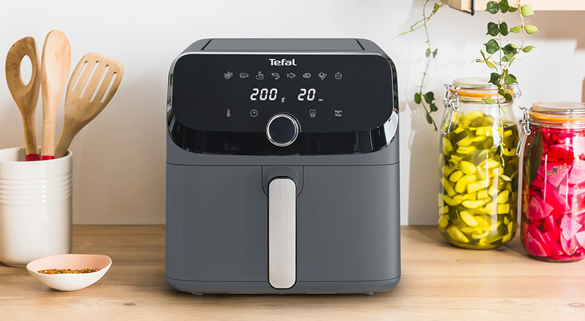 Ein grauer Tefal Airfryer steht auf einem Holztisch neben Küchenutensilien und eingelegten Gurken und Rüben.