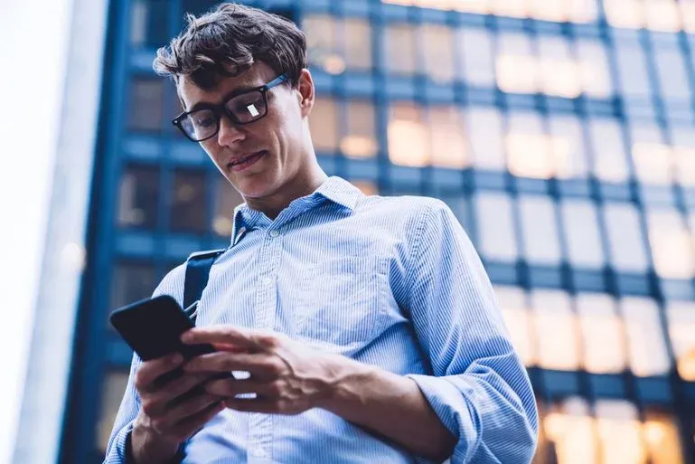 Eine Person mit Brille und blau gestreiftem Hemd tippt auf ein schwarzes Smartphone, das sie in beiden Händen hält.