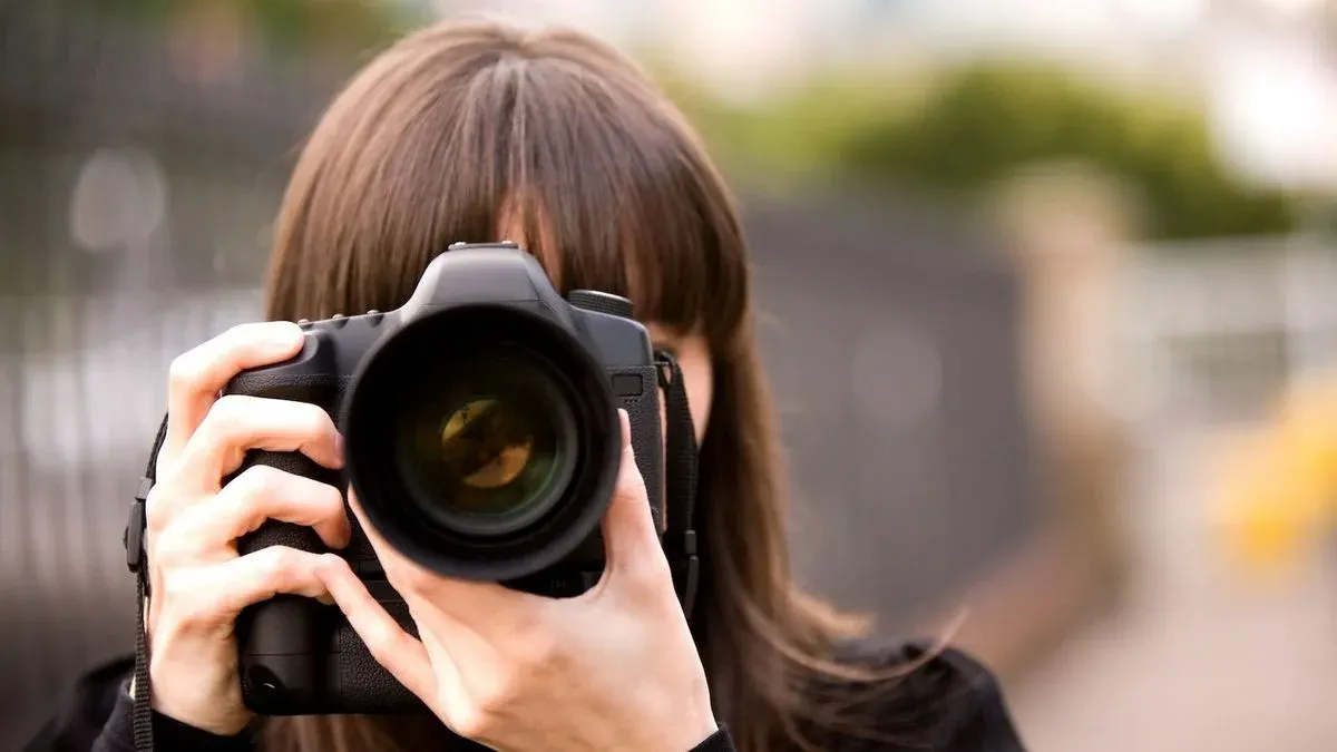 Person mit Haaren vor dem Gesicht hält schwarze DSLR-Kamera mit großem Objektiv. Unscharfer Hintergrund in Grün- und Grautönen.