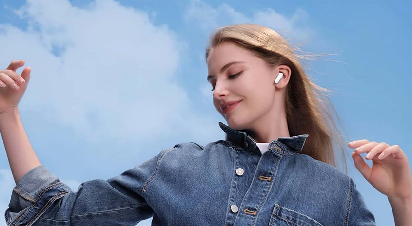 Eine Frau mit einem weißen Ohrhörer und einer blauen Jeansjacke vor einem bewölkten, blauen Himmel.