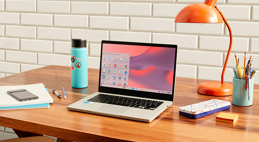 Ein Holzschreibtisch mit Laptop, Lampe, Wasserflasche, Stiften, Notizen und einem Stifthalter vor einer weißen Ziegelwand.