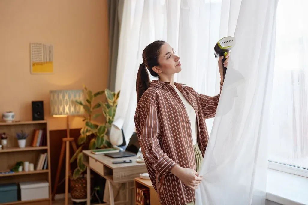 Eine Person in einem gestreiften Hemd dämpft weiße Vorhänge mit einem Handdampfglätter. Bücherregal, Lampe, Pflanze und Schreibtisch im Hintergrund.