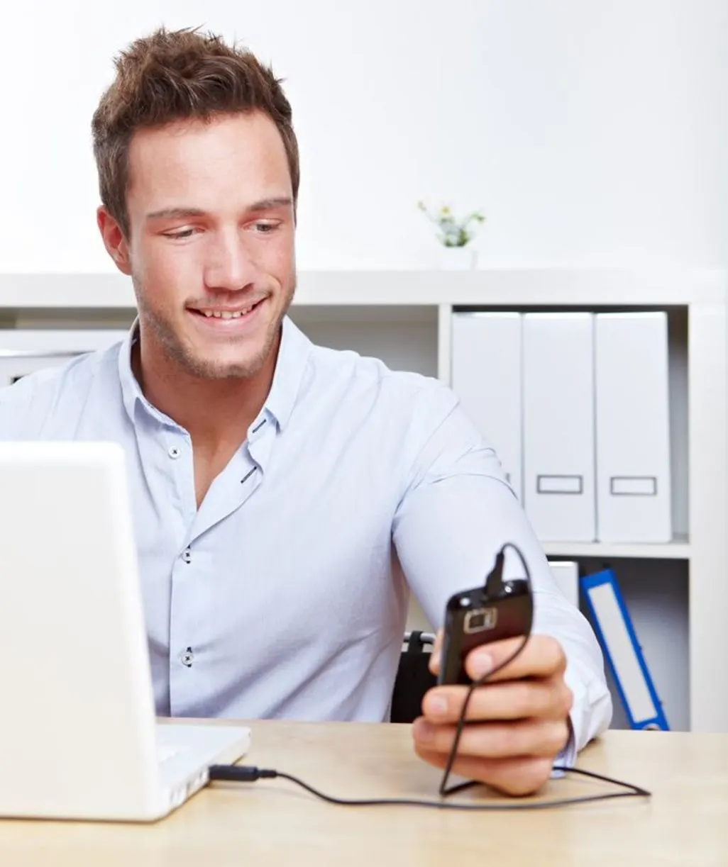 Mann sitzt am Schreibtisch vor einem Laptop und hält ein Smartphone in der Hand.