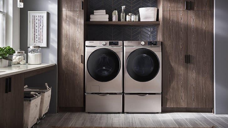 Zwei Waschmaschinen auf Podesten in einem Raum mit dunklen Holzschränken und Arbeitsplatte. Dunkle Fliesenwand mit Regal, Gläsern und Pflanzen.