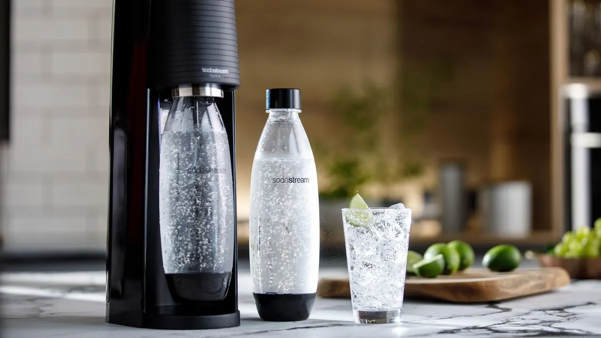 Schwarzes SodaStream Gerät mit Sprudelflasche, weitere Flasche und Glas mit Limette auf Marmorplatte. Limetten und Trauben im Hintergrund.