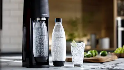 Schwarzes SodaStream Gerät mit Sprudelflasche, weitere Flasche und Glas mit Limette auf Marmorplatte. Limetten und Trauben im Hintergrund.