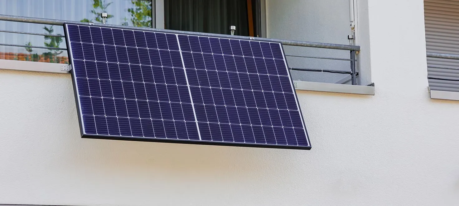 Ein Solarmodul hängt an einem Balkongeländer vor einer weißen Wand mit Fenstern.