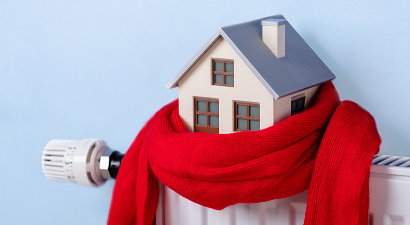 Ein Hausmodell mit rotem Schal auf einem Heizkörper vor blauem Hintergrund.