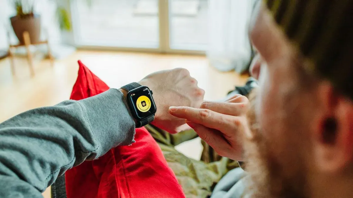 Eine Person mit grauem Oberteil tippt auf eine schwarze Smartwatch mit gelbem Kamerasymbol. Roter Stoff und ein Fenster im Hintergrund.