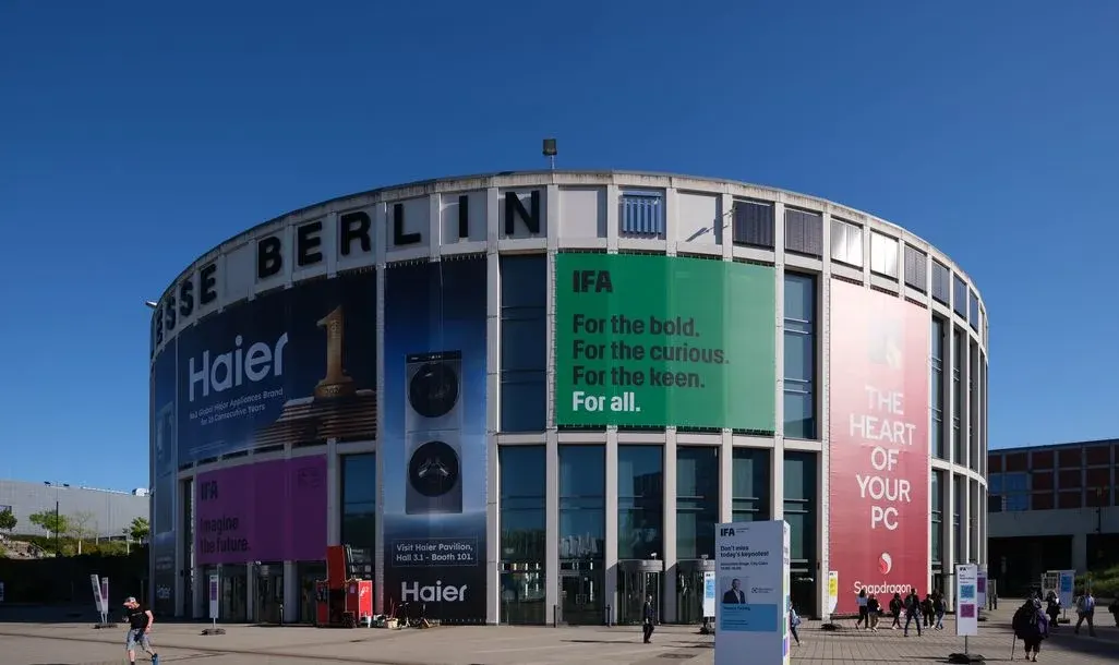 Gekrümmtes Messegebäude "MESSE BERLIN" mit großen blauen, grünen, rosa und roten IFA-Bannern. Menschen auf gepflastertem Platz unter blauem Himmel.