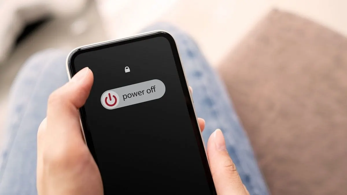 Eine Hand hält ein schwarzes Smartphone mit „power off“-Anzeige. Im Hintergrund sind verschwommene blaue Jeans und eine braune Oberfläche zu sehen.
