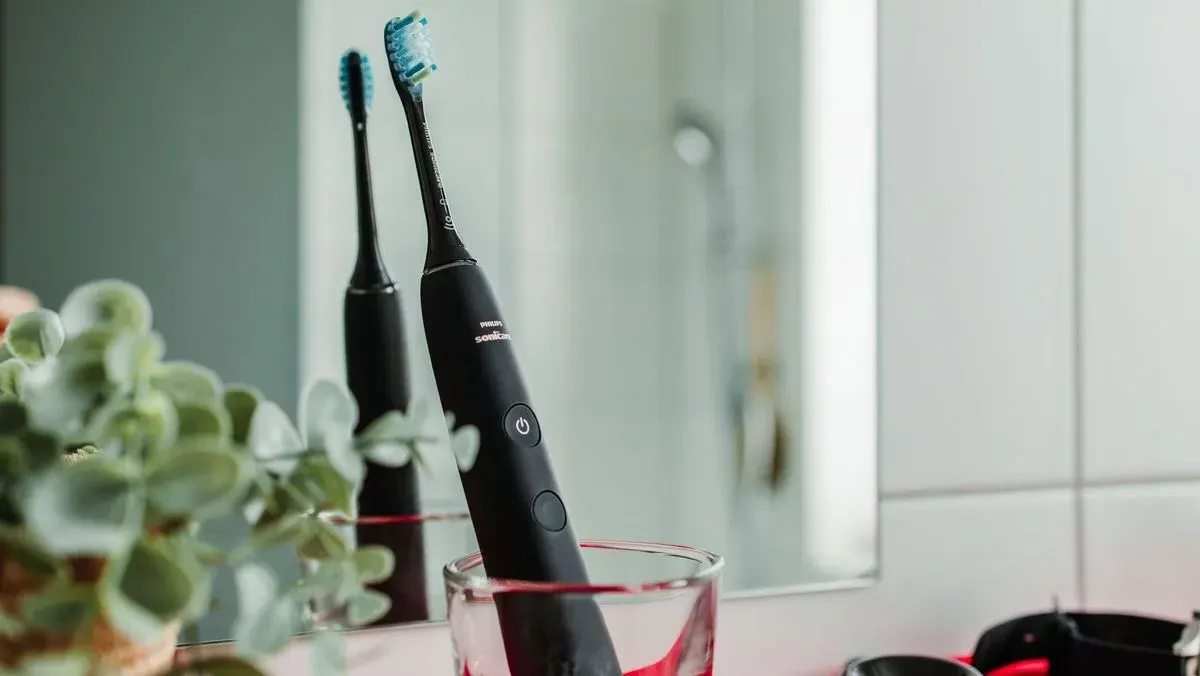 Schwarze Philips Sonicare Elektrozahnbürste mit blau-grünem Bürstenkopf in einem Glas. Grüne Pflanze, Spiegel und weiße Wand.