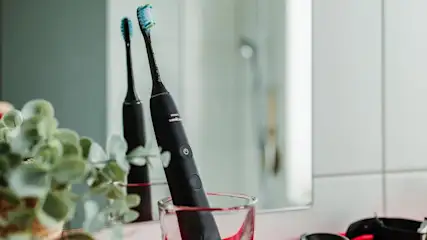 Schwarze Philips Sonicare Elektrozahnbürste mit blau-grünem Bürstenkopf in einem Glas. Grüne Pflanze, Spiegel und weiße Wand.