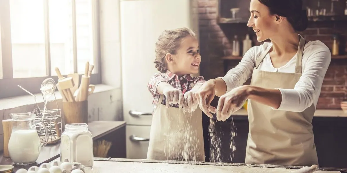 Erwachsene Person und Kind streuen gemeinsam Mehl auf eine Arbeitsfläche in der Küche.