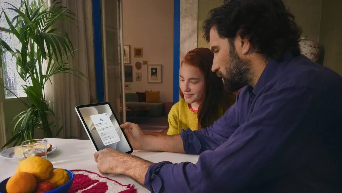 Vater sitzt mit Tochter an einem Tisch und beide sehen sich Inhalte auf einem iPad an.
