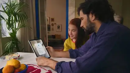 Vater sitzt mit Tochter an einem Tisch und beide sehen sich Inhalte auf einem iPad an.