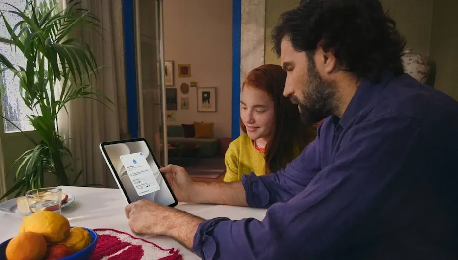Vater sitzt mit Tochter an einem Tisch und beide sehen sich Inhalte auf einem iPad an.