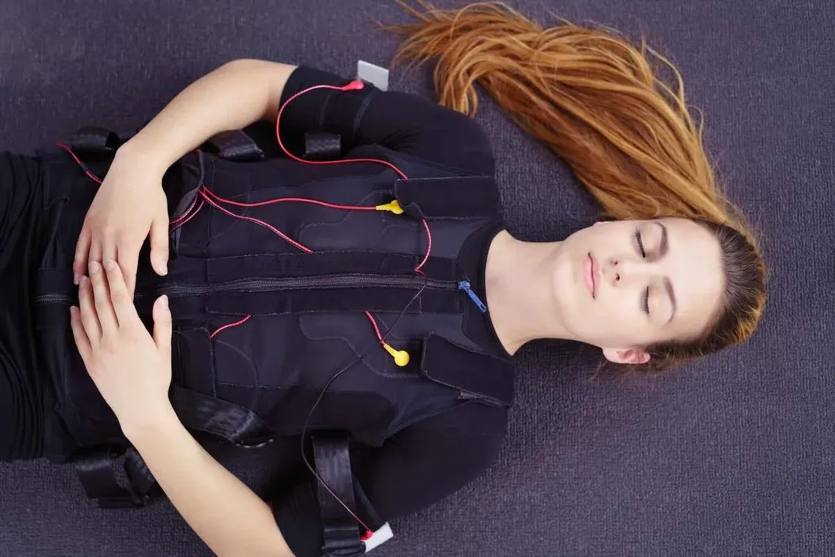 Frau in EMS‑Anzug liegt entspannt auf dem Rücken auf einer Matte, mit geschlossenen Augen und gefalteten Händen auf dem Bauch.