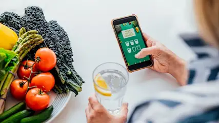 Person hält ein Glas Wasser mit Zitronenscheibe in der einen Hand und benutzt mit der anderen Hand ein Smartphone mit einer App zur Überwachung des Wasserhaushalts.