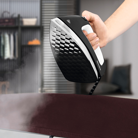 Eine Hand hält ein schwarzes Bügeleisen mit weißem Griff über ein braunes Brett. Im Hintergrund ein Schrank.