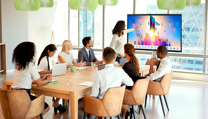 Eine Gruppe von Leuten sitzt an einem Holztisch und schaut auf einen großen Bildschirm in einem Konferenzraum.