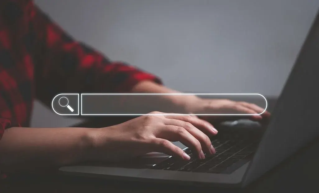 Hände tippen auf Laptop‑Tastatur, darüber eine eingeblendete Suchleiste mit Lupen‑Symbol.