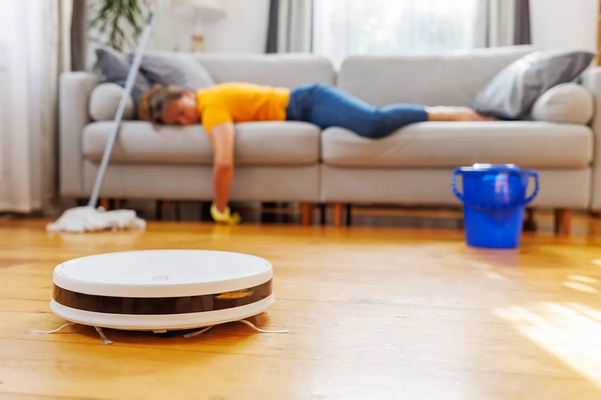 Weißer Saugroboter vor einer Couch, auf der eine müde Person liegt. Vor der Couch sind ein Wischmopp und ein blauer Kübel zu sehen.