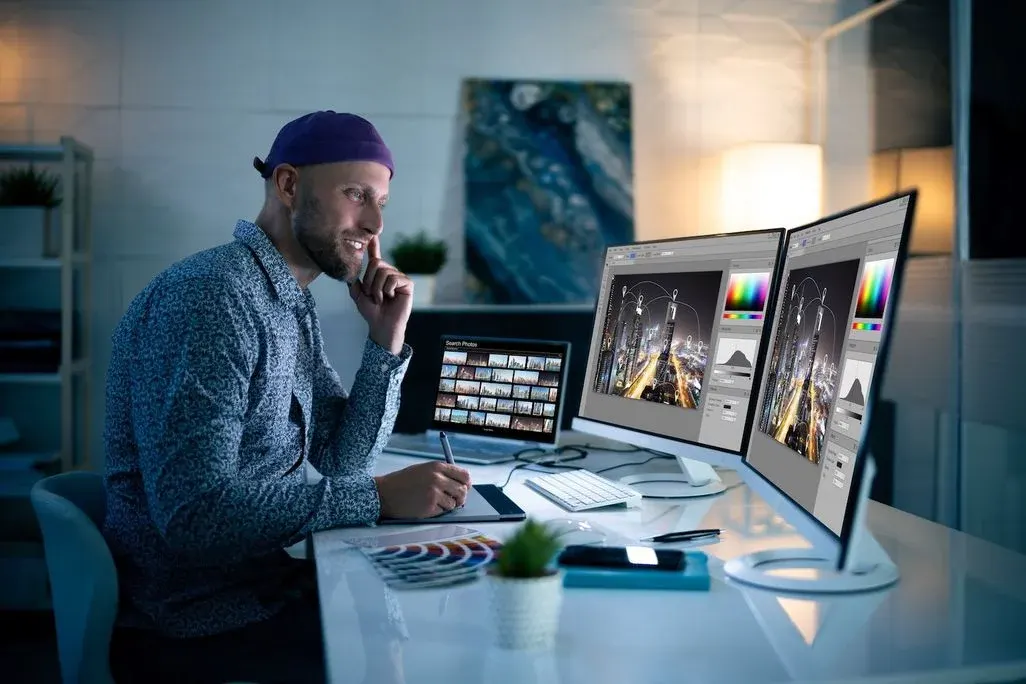 ein Mann sitzt am Schreibtisch, vor sich 3 Bildschirme, er bearbeitet Fotos