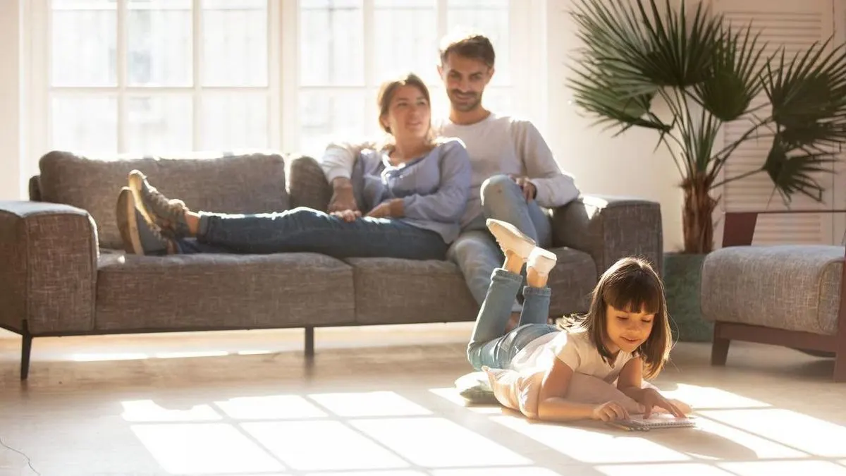 Kind zeichnet auf dem Boden, während zwei Erwachsene auf einem grauen Sofa sitzen. Heller Raum mit großem Fenster und Pflanze.