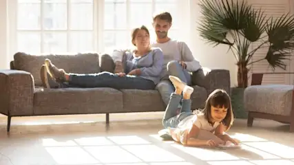 Kind zeichnet auf dem Boden, während zwei Erwachsene auf einem grauen Sofa sitzen. Heller Raum mit großem Fenster und Pflanze.