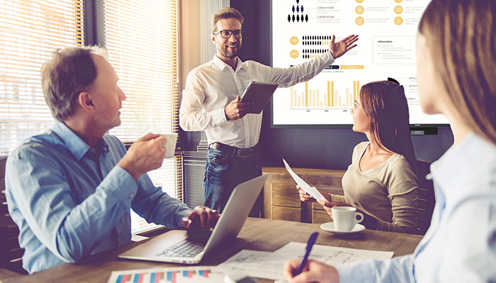 Eine Bürobesprechung mit Diagrammen auf einem Bildschirm. Ein Mann präsentiert, andere sitzen mit Kaffee und Laptop am Tisch.