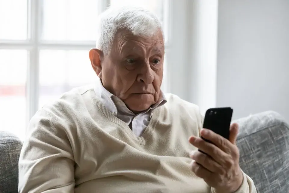 Ältere Person in hellem Pullover blickt überrascht auf ein schwarzes Smartphone in ihrer Hand.
