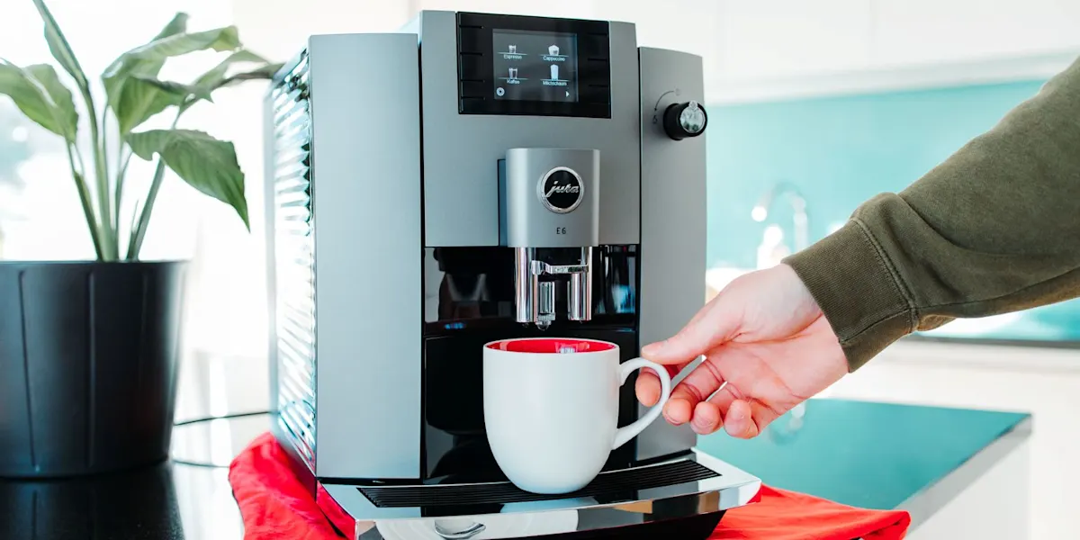 Graue Jura E6 Kaffeemaschine mit Display. Eine Hand platziert eine weiß-rote Tasse auf schwarzer Arbeitsplatte. Rotes Tuch, Pflanze, blaue Wand.