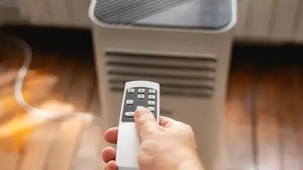 Hand mit Fernbedienung zeigt auf mobiles Klimagerät, das auf einem Holzboden in der Sonne vor einem Fenster steht.