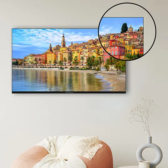 Ein Flachbildfernseher an einer Wand zeigt eine Küstenstadt mit farbenfrohen Häusern, Palmen und einem Strand unter blauem Himmel.