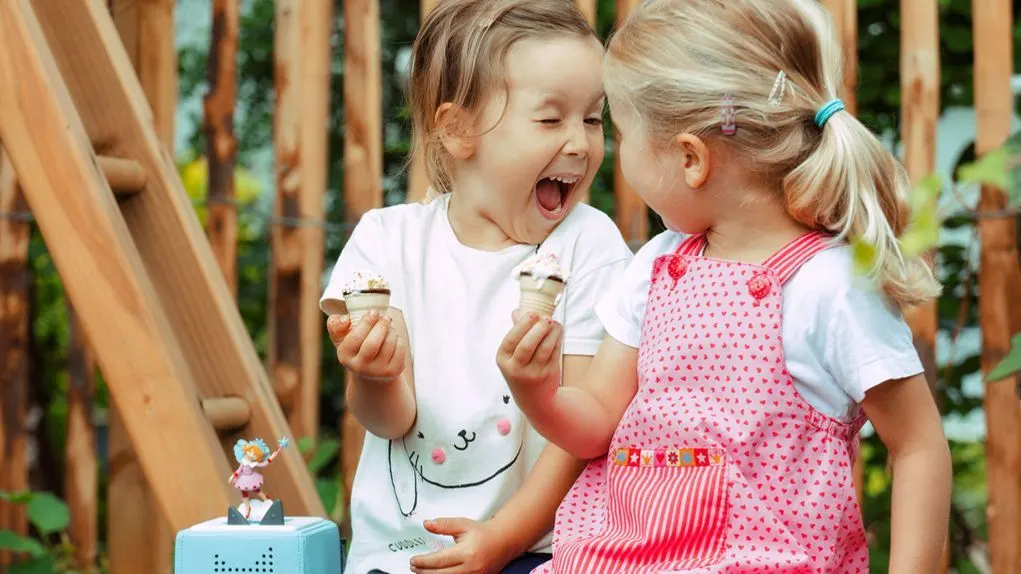 2 Kinder sitzen auf einer Holzrutsche und essen Eiswaffeln, vor ihnen steht eine blaue Toniebox