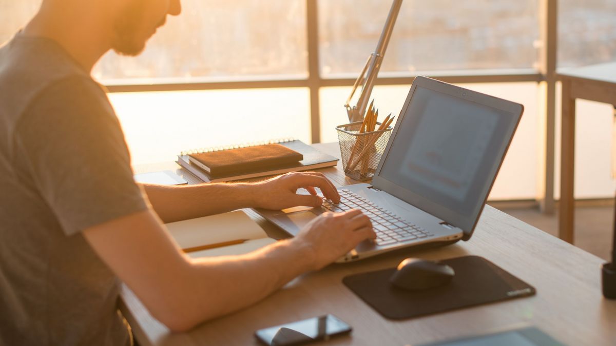 Person arbeitet am Laptop bei Sonnenlicht mit Notizbüchern und Stiften am Schreibtisch.