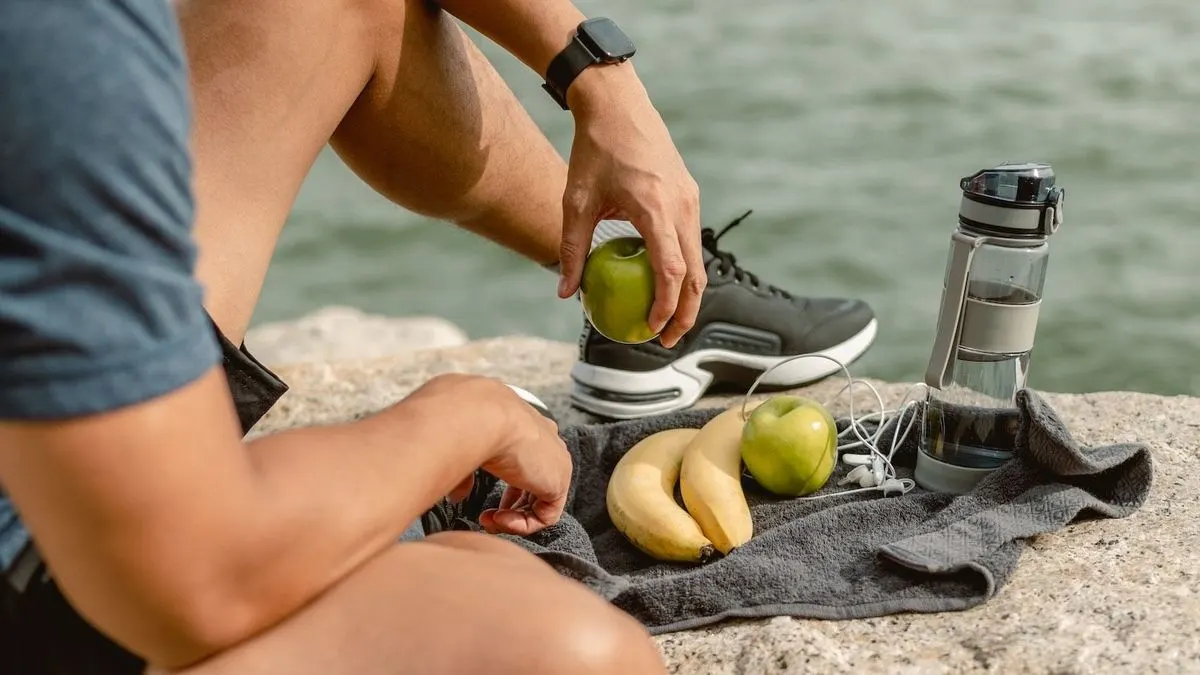 Eine Person sitzt an einer Klippe mit Blick auf Wasser und hält einen Apfel, vor ihm liegt ein weiterer Apfel, Bananen und eine Wasserflasche.