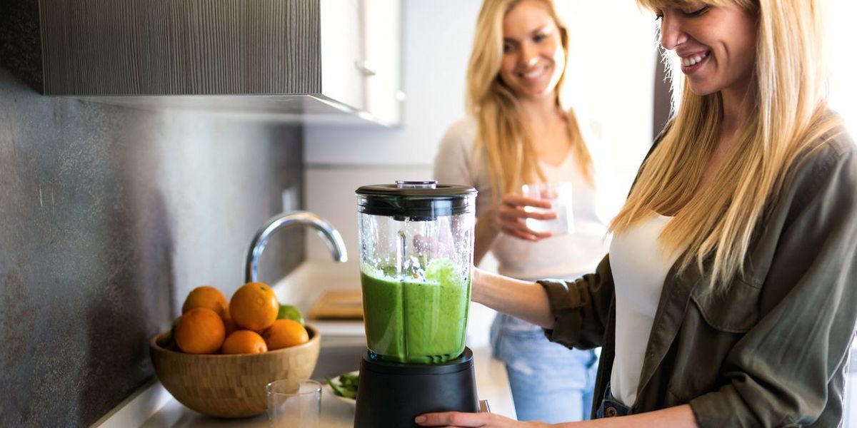 Zwei Frauen in der Küche, eine mixt einen grünen Smoothie, die andere hält ein Glas.