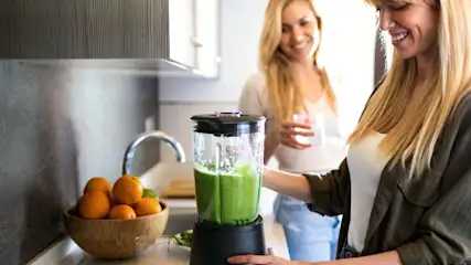 Zwei Frauen in der Küche, eine mixt einen grünen Smoothie, die andere hält ein Glas.