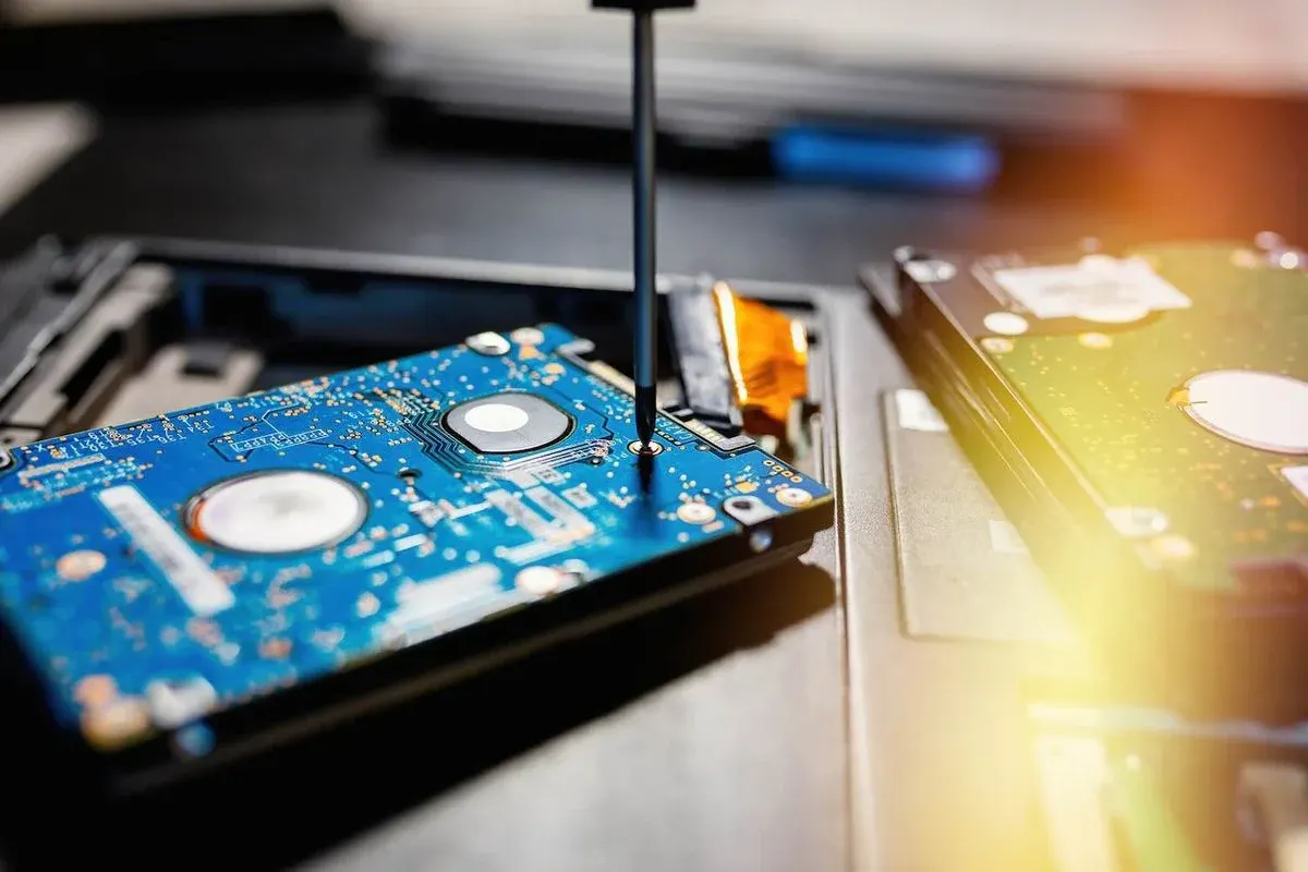Nahaufnahme einer geöffneten Festplatte im Laptop, auf deren blauer Platine ein Schraubendreher ansetzt; daneben weitere Hardwarekomponenten unscharf.